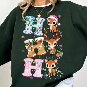 Könnte beinhalten: Dunkelgrünes Sweatshirt mit dem Schriftzug "HO HO HO" in festlichen Buchstaben. Die Buchstaben sind mit Weihnachtsbeleuchtung und Mustern verziert. Niedliche Cartoon-Hirsche mit Weihnachtsmützen und Zuckerstangen sind ebenfalls abgebildet.