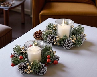 May include: Two lit candles in clear glass holders, surrounded by a festive arrangement of pine needles, pine cones, red berries, and gold-colored beads. The candles are on a light gray surface, suggesting a holiday or seasonal theme.