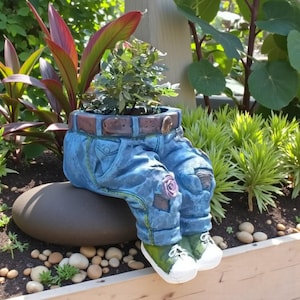 Puede incluir: Una maceta caprichosa de mezclilla azul con un cinturón marrón y una pequeña planta en el interior. La maceta tiene piernas con zapatillas verdes y blancas. Un elemento decorativo de jardín.