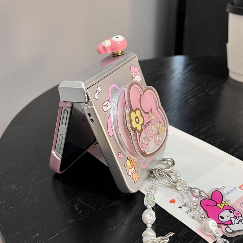 May include: Silver and pink foldable phone with a My Melody design. The phone features a pink pop socket and a small pink charm on top. A beaded keychain with a My Melody charm hangs from the phone.