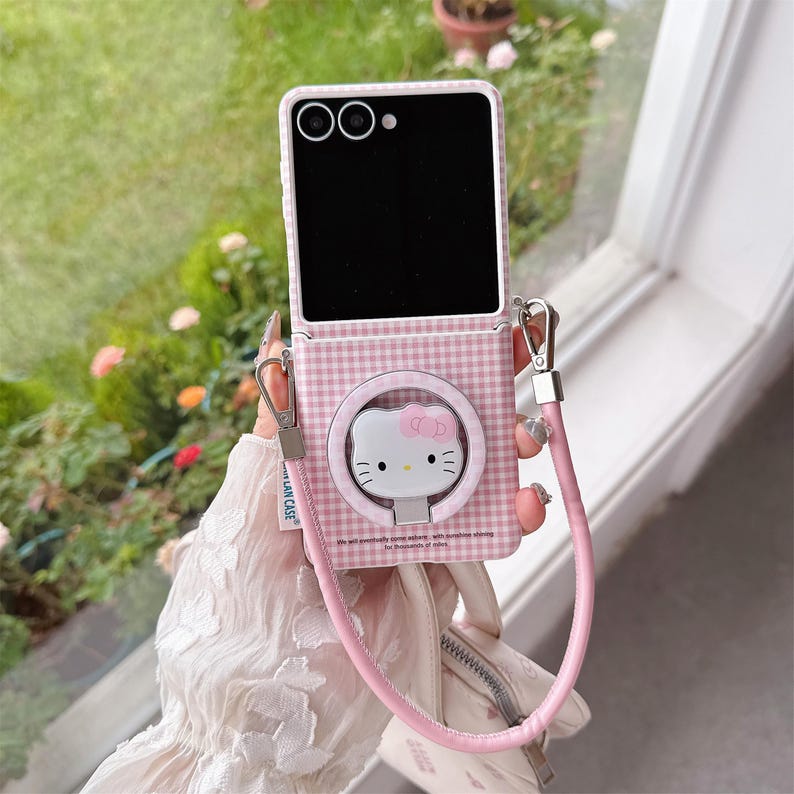 May include: Pink and white checked phone case with a Hello Kitty design. The case has a pink strap and silver hardware. The phone screen is black. The case has the text: "We will passionately love others, with sunshine shining for thousands of miles."
