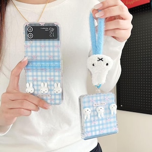 May include: Clear phone case featuring a blue gingham design, pink bows, and white bunny and tooth-shaped decorations. The case has the text "Be happy every day". A matching plush charm is attached to a blue strap.