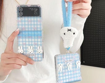 May include: Clear phone case with a blue gingham pattern, pink bows, and white bunny and tooth-shaped embellishments. The case has the text "Be happy every day". A matching plush charm is attached to a blue strap.