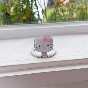 May include: A silver phone ring holder with a Hello Kitty design. The holder has a white face with a pink bow, black whiskers, and a silver ring for grip. The ring is attached to a white base.