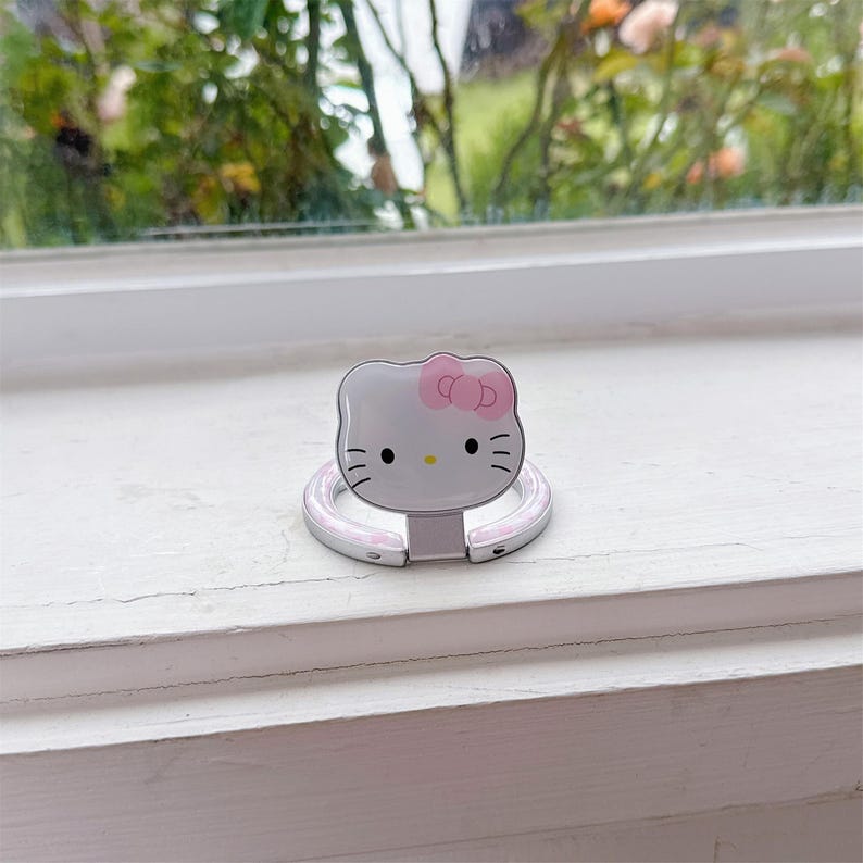 May include: A silver phone ring holder with a Hello Kitty design. The holder has a white face with a pink bow, black whiskers, and a silver ring for grip. The ring is attached to a white base.