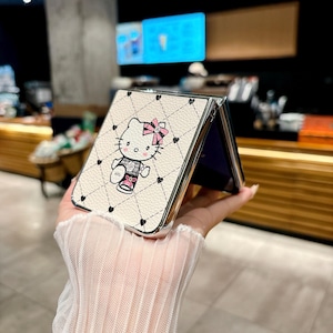 May include: A white phone case with a quilted pattern and a cartoon image of Hello Kitty wearing a pink bow. The case has a silver frame and black heart accents. The phone case is open, revealing a purple interior.