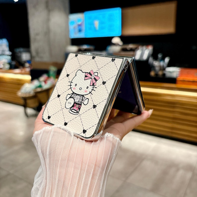 May include: A white phone case with a quilted pattern and a cartoon image of Hello Kitty wearing a pink bow. The case has a silver frame and black heart accents. The phone case is open, revealing a purple interior.