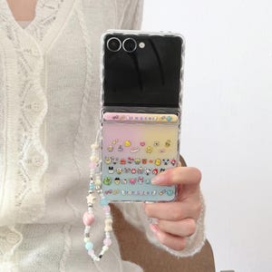 May include: Clear phone case with a colorful gradient design and a variety of cartoon character stickers. The case has a beaded wrist strap with pastel-colored beads and star-shaped charms. The phone case is open, revealing the phone's camera.