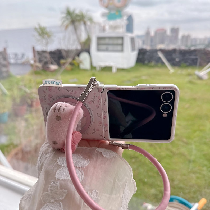 May include: A pink phone case with a floral pattern and a matching pink strap. The case has a small plush character attached. The phone screen is black. The text "LAN LAN CASE" is visible on the case.