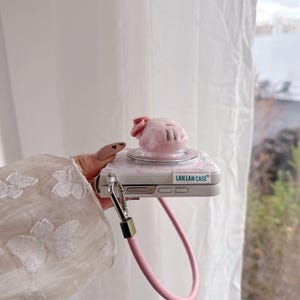 May include: A pink phone case with a plush pig-shaped charm and a pink strap. The case has a floral design and the text "LAN LAN CASE". The charm has a small pink bow.