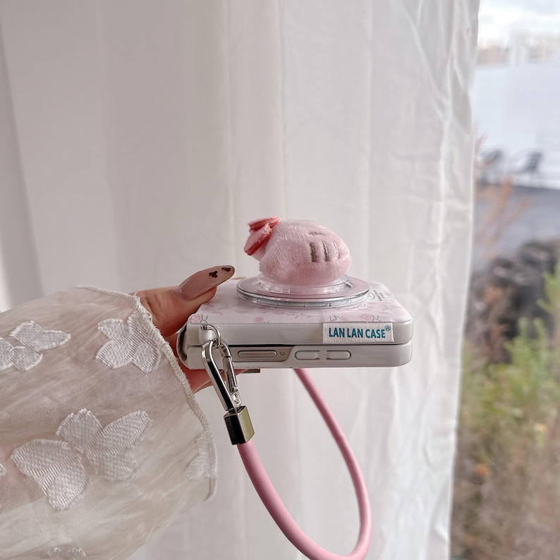 May include: A pink phone case with a plush pig-shaped charm and a pink strap. The case has a floral design and the text "LAN LAN CASE". The charm has a small pink bow.