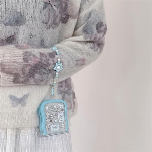 May include: A light blue, handheld, clear plastic case with a cartoon design is attached to a beaded bracelet with a star-shaped charm. The case is hanging from a light blue and white beaded bracelet. The person is wearing a light blue sweater with a floral pattern.