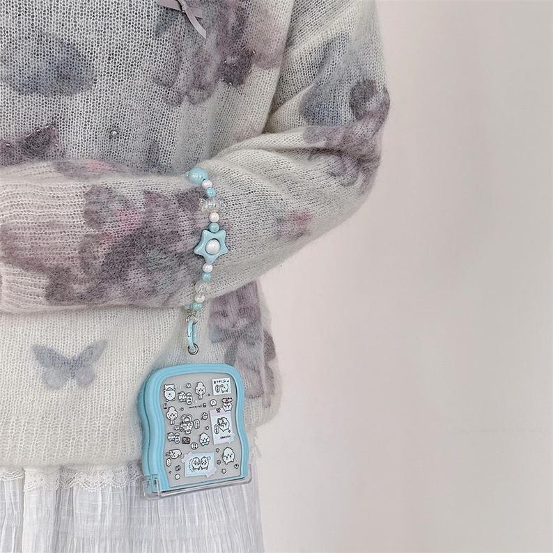 May include: A light blue, handheld, clear plastic case with a cartoon design is attached to a beaded bracelet with a star-shaped charm. The case is hanging from a light blue and white beaded bracelet. The person is wearing a light blue sweater with a floral pattern.