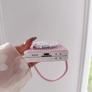May include: A pink and white gingham patterned phone case with a matching pop-up grip. The case has a white border and a pink strap. The pop-up grip is silver and white.