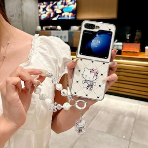 May include: A white flip phone case with a Hello Kitty design, featuring a pink bow and black heart accents. The case has a diamond pattern and a matching charm bracelet with pearl and crystal beads.