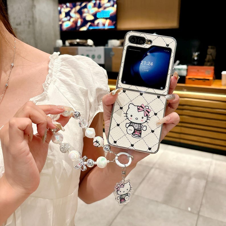 May include: A white flip phone case with a Hello Kitty design, featuring a pink bow and black heart accents. The case has a diamond pattern and a matching charm bracelet with pearl and crystal beads.