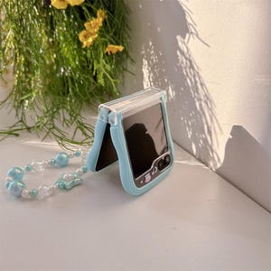 May include: A light blue phone case with a clear top, designed for a Samsung phone. The case has a matching beaded wrist strap with light blue and white beads. The phone screen is black.