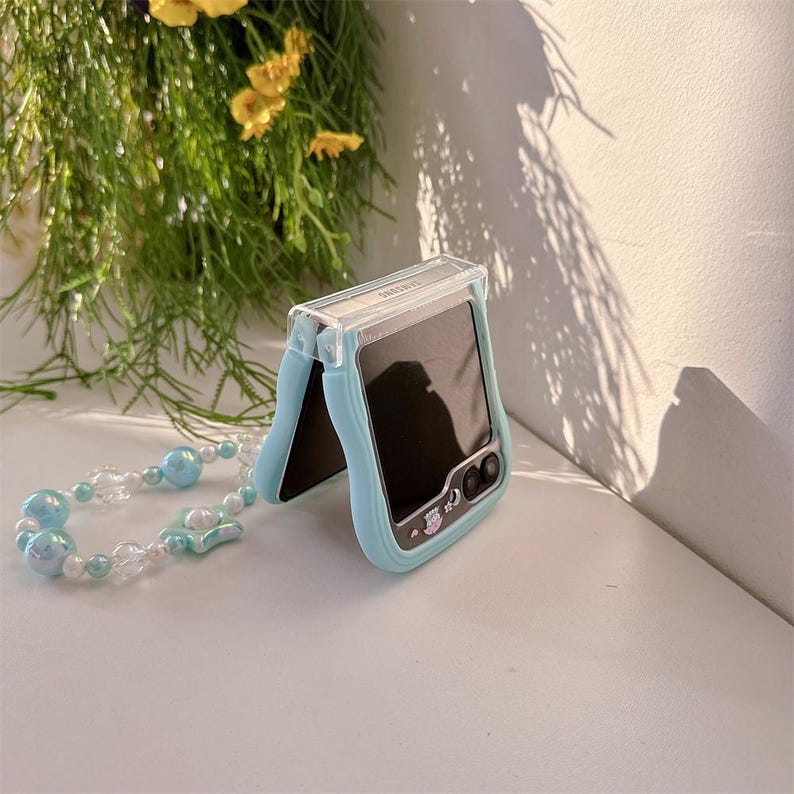 May include: A light blue phone case with a clear top, designed for a Samsung phone. The case has a matching beaded wrist strap with light blue and white beads. The phone screen is black.