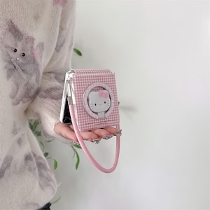 May include: Pink and white gingham phone case with a Hello Kitty design. The case has a circular ring holder and a pink strap. The case is held in front of a person wearing a white sweater with a floral design.
