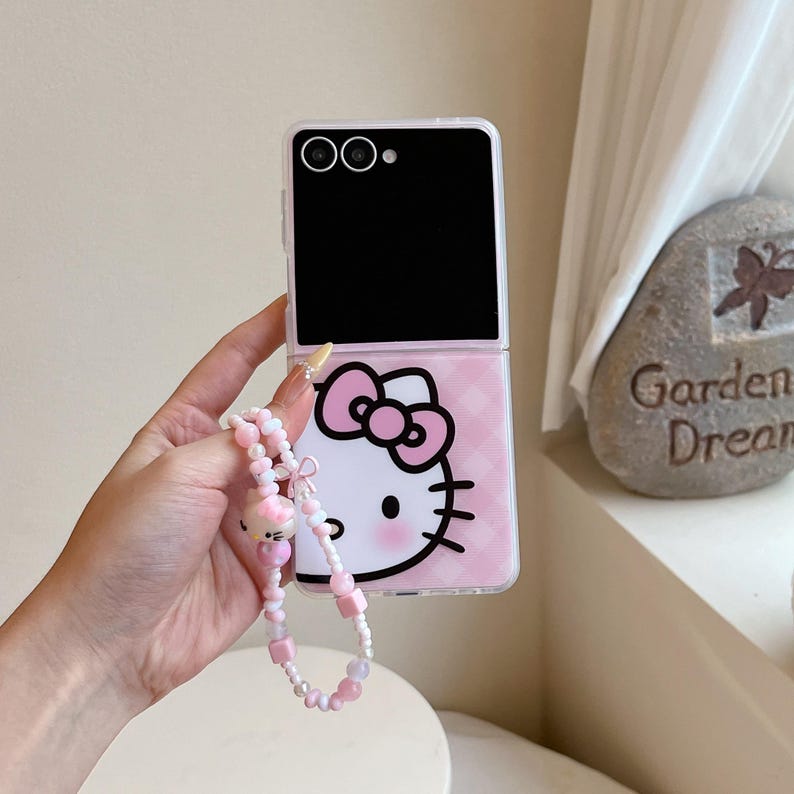 May include: A pink and white phone case with a Hello Kitty design. The case has a black top section and a pink checkered pattern on the bottom half. A beaded wrist strap with pink and white beads and a Hello Kitty charm is attached.