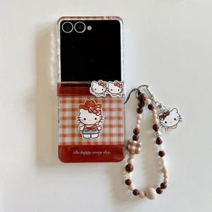 Op de afbeelding: Transparante telefoonhoes met een rood en wit ruitpatroon, met Hello Kitty-afbeeldingen. De hoes heeft een bijpassende bedel met een kralenband en een Hello Kitty-bedel. De tekst "Be happy every day" is op de hoes gedrukt.
