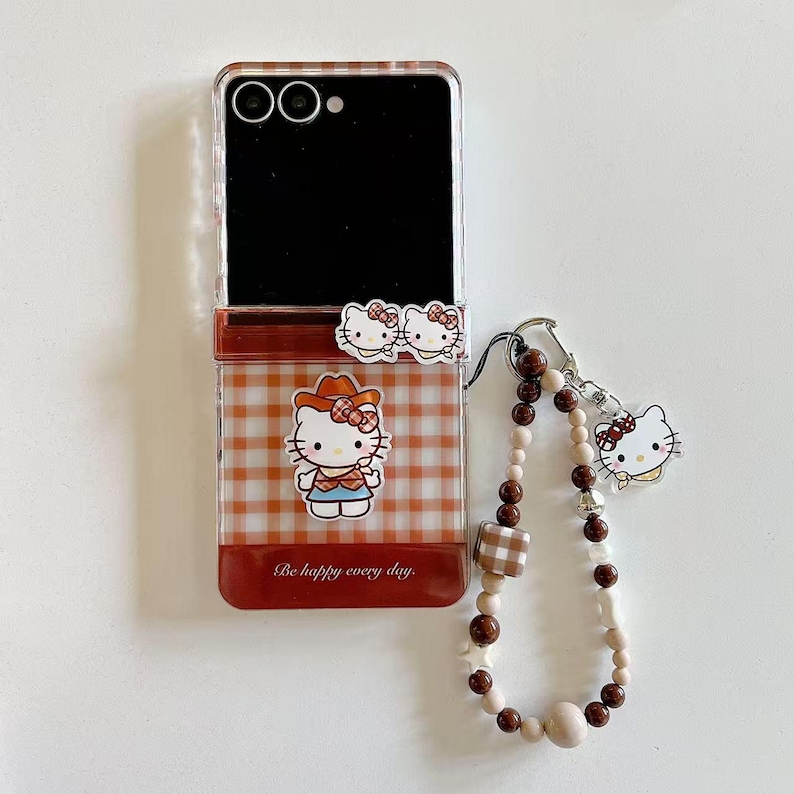 Op de afbeelding: Transparante telefoonhoes met een rood en wit ruitpatroon, met Hello Kitty-afbeeldingen. De hoes heeft een bijpassende bedel met een kralenband en een Hello Kitty-bedel. De tekst "Be happy every day" is op de hoes gedrukt.