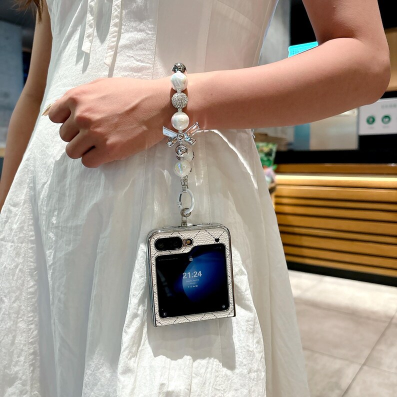 May include: A silver phone case with a white quilted pattern is attached to a pearl and rhinestone bracelet. The phone screen displays the time 21:24. The bracelet is connected to the phone case with a silver chain.