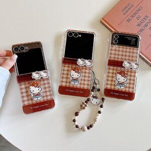 Op de afbeelding: Drie transparante telefoonhoesjes met een rood-wit geruit patroon, versierd met een Hello Kitty-afbeelding. Elke hoes heeft een rode band en een Hello Kitty-bedel. Er is ook een kralen polsbandje met een Hello Kitty-bedel. De tekst "Be happy every day" is zichtbaar.