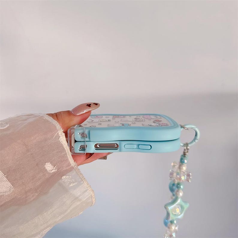 May include: A light blue phone case with a cartoon design. The case has a clear border and a decorative strap with beads and a star-shaped charm. The phone case is held by a person.