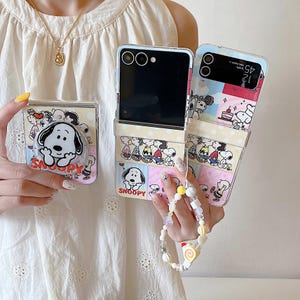 May include: Clear phone cases featuring Snoopy and other Peanuts characters. The cases have a pop socket and a wrist strap with beads. The cases are designed to fit a foldable phone. The word "Snoopy" is printed on the cases.