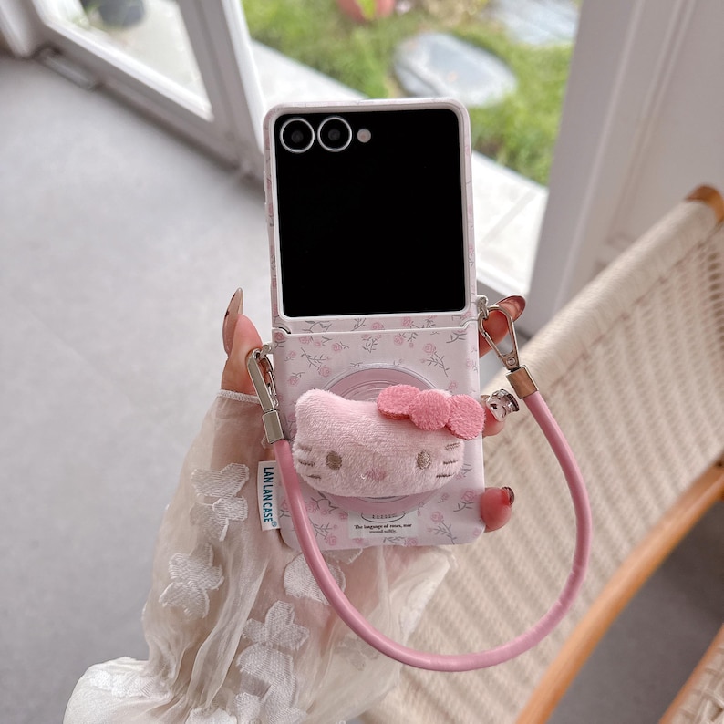 May include: A pink phone case with a floral pattern and a plush Hello Kitty face. The case has a matching pink strap and silver-colored clasps. The phone screen is black.