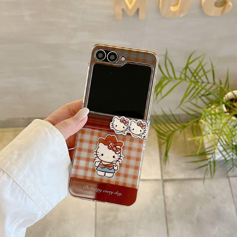 Op de afbeelding: Transparante telefoonhoes met een rood-bruin ruitpatroon en Hello Kitty-afbeeldingen. De hoes heeft een scharnierend ontwerp en bevat de tekst "Be happy every day."