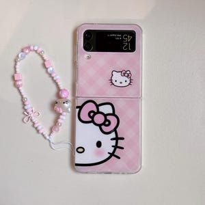 May include: A pink phone case with a Hello Kitty design, and a matching beaded wrist strap. The case has a gingham pattern and a clear plastic overlay. The wrist strap includes pink and white beads and a bow.