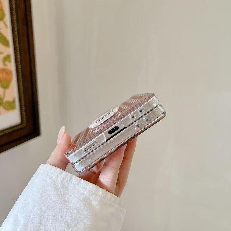 Op de afbeelding: Een doorzichtige telefoonhoes met een roze en wit gestreept ontwerp. De hoes heeft een ingebouwde ringhouder en is gemaakt van transparant materiaal. De telefoonhoes is ontworpen om de telefoon te beschermen.