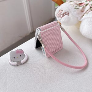 May include: A pink and white gingham phone case with a matching pink strap and a silver clasp. A silver phone ring with a Hello Kitty design is also present. The phone case is designed to fit a foldable phone.