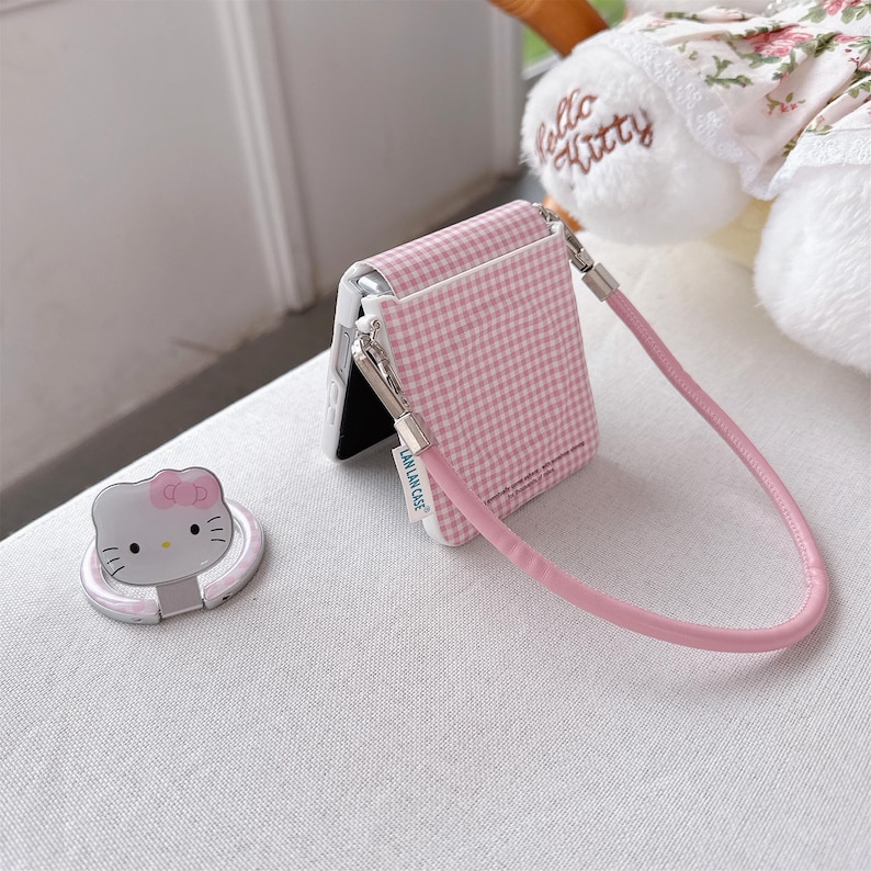 May include: A pink and white gingham phone case with a matching pink strap and a silver clasp. A silver phone ring with a Hello Kitty design is also present. The phone case is designed to fit a foldable phone.