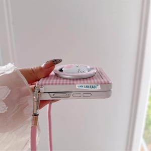 May include: A pink and white gingham phone case with a matching pop socket featuring a cat design. The case has a silver frame and a pink strap. The text "LAN LAN CASE" is printed on the case.