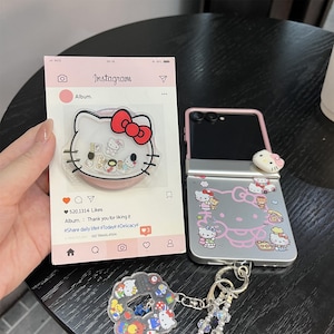 May include: A collection of Hello Kitty themed accessories. Includes a phone case, a phone grip, and a keychain. The phone case is silver with pink accents and Hello Kitty designs. The phone grip is clear with a pink base and a Hello Kitty design. The keychain has a Hello Kitty charm.