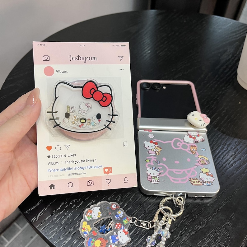 May include: A collection of Hello Kitty themed accessories. Includes a phone case, a phone grip, and a keychain. The phone case is silver with pink accents and Hello Kitty designs. The phone grip is clear with a pink base and a Hello Kitty design. The keychain has a Hello Kitty charm.