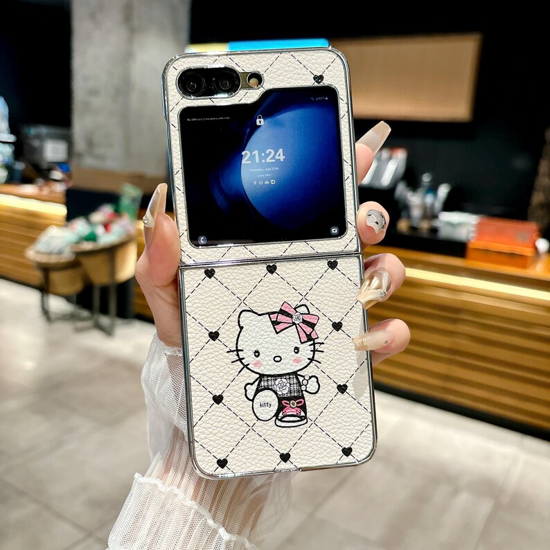 May include: White, foldable phone case featuring a Hello Kitty design with a pink bow and black heart accents. The case has a quilted pattern and a silver trim. The phone screen displays the time 21:24.