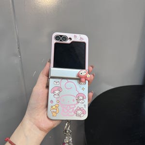 May include: A pink and silver flip phone case featuring a cartoon character design. The case has a pink top and silver bottom with a cartoon character and other graphics. A small charm hangs from the bottom of the case.