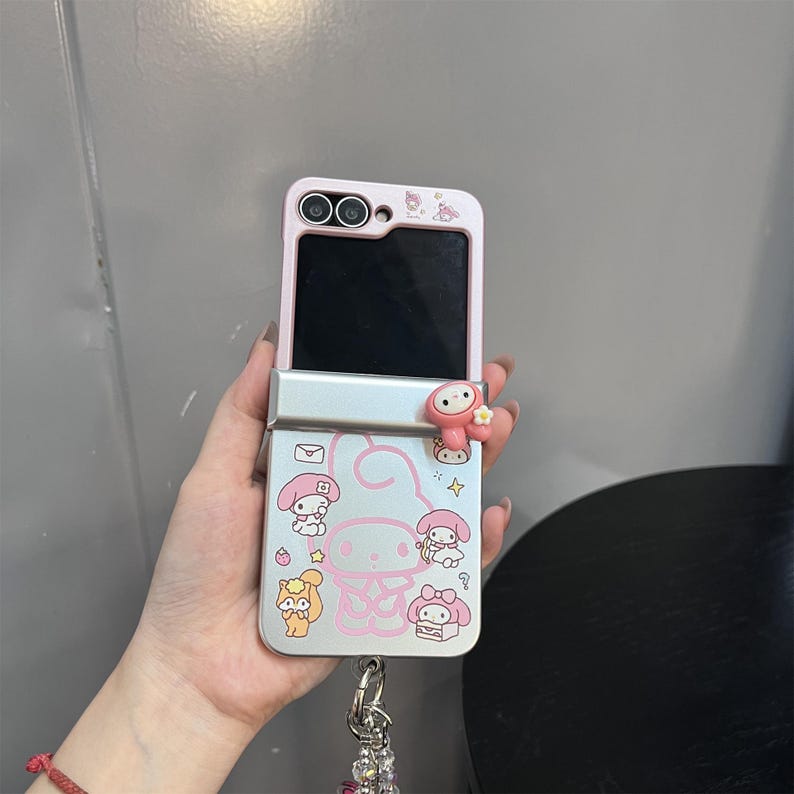 May include: A pink and silver flip phone case featuring a cartoon character design. The case has a pink top and silver bottom with a cartoon character and other graphics. A small charm hangs from the bottom of the case.