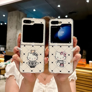 May include: Two white flip phone cases with a quilted pattern and Hello Kitty designs. The cases feature black hearts and a cartoon Hello Kitty character with a bow and outfit. The phone screens are visible, displaying the time.