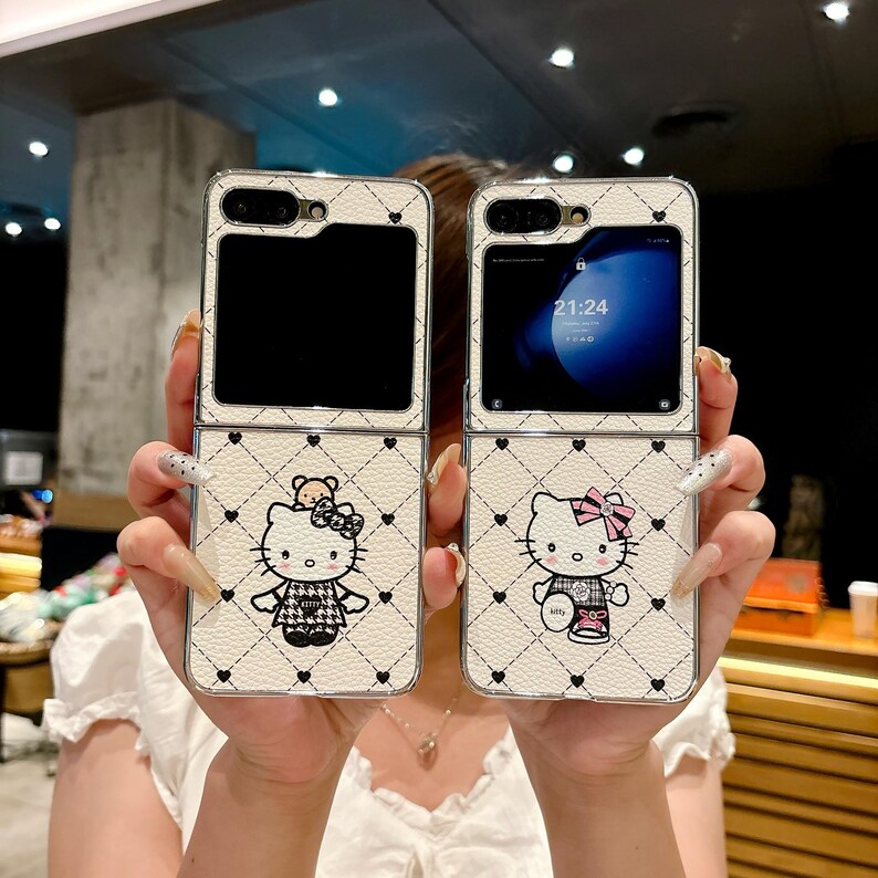 May include: Two white flip phone cases with a quilted pattern and Hello Kitty designs. The cases feature black hearts and a cartoon Hello Kitty character with a bow and outfit. The phone screens are visible, displaying the time.