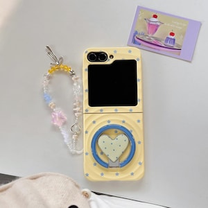 May include: A yellow phone case featuring a heart-shaped grip and star detailing. It has a beaded wrist strap with colourful beads and charms. A small card with a dessert illustration is also visible.