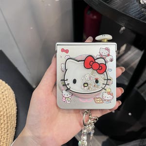 May include: A silver-colored compact mirror featuring a Hello Kitty design. The mirror has a clear, plastic overlay with a white Hello Kitty face, a red bow, and other character illustrations. A beaded keychain is attached.