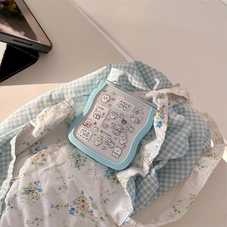 May include: A light blue, wavy-edged case with various cartoon character stickers. The case rests on a blue and white floral patterned fabric inside a blue and white checkered tote bag. A black tablet is visible in the background.