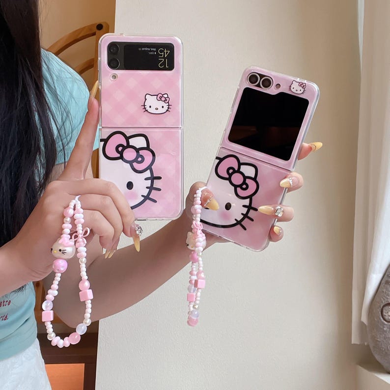 May include: Two pink flip phone cases with Hello Kitty designs. The cases have a pink checkered pattern and a Hello Kitty graphic. Each case has a matching pink and white beaded wrist strap with a Hello Kitty charm.