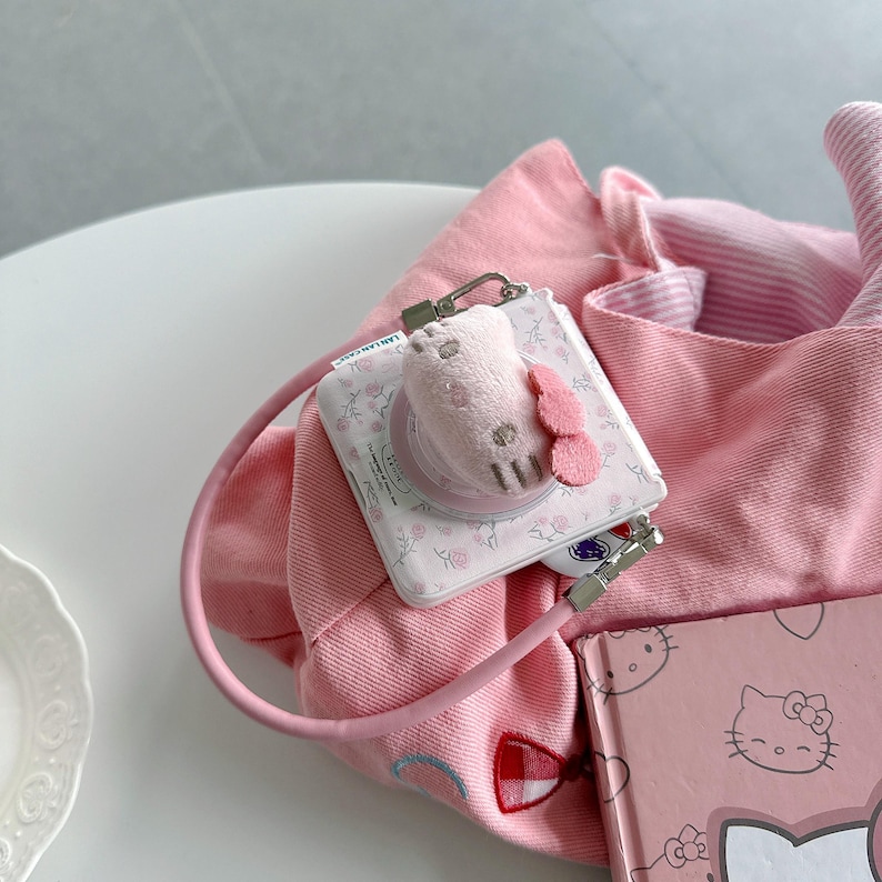 May include: A pink tote bag with a matching phone grip featuring a plush Hello Kitty design. The phone grip is attached to a white floral-patterned phone stand. A small pink notebook with a Hello Kitty illustration is also visible.