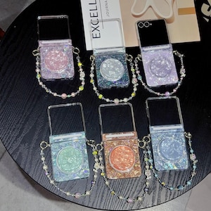 May include: Six foldable phone cases with glittery, iridescent shell-like textures in pink, silver, purple, green, orange, and blue. Each case features a circular pop-out grip and a decorative beaded wrist strap with small charms and pearls.